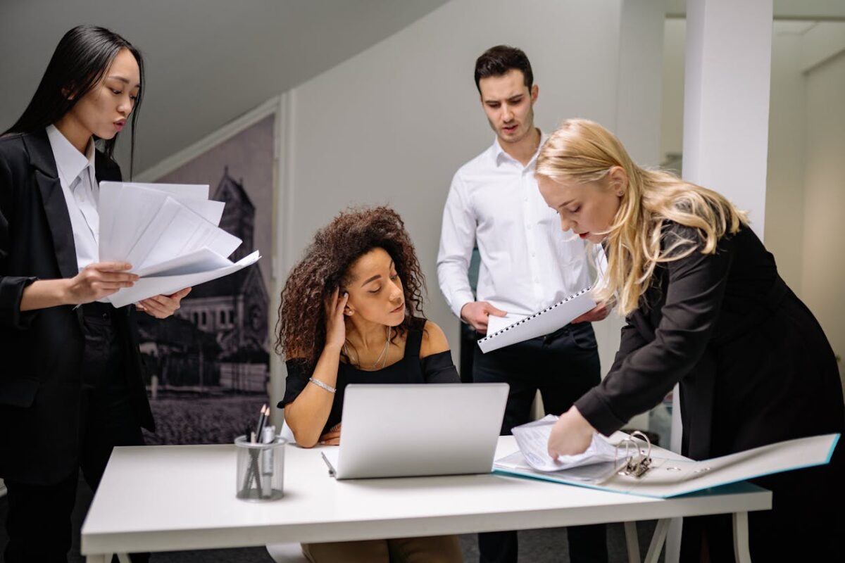 A diverse group of coworkers brainstorming over paperwork and laptops in a corporate office setting.